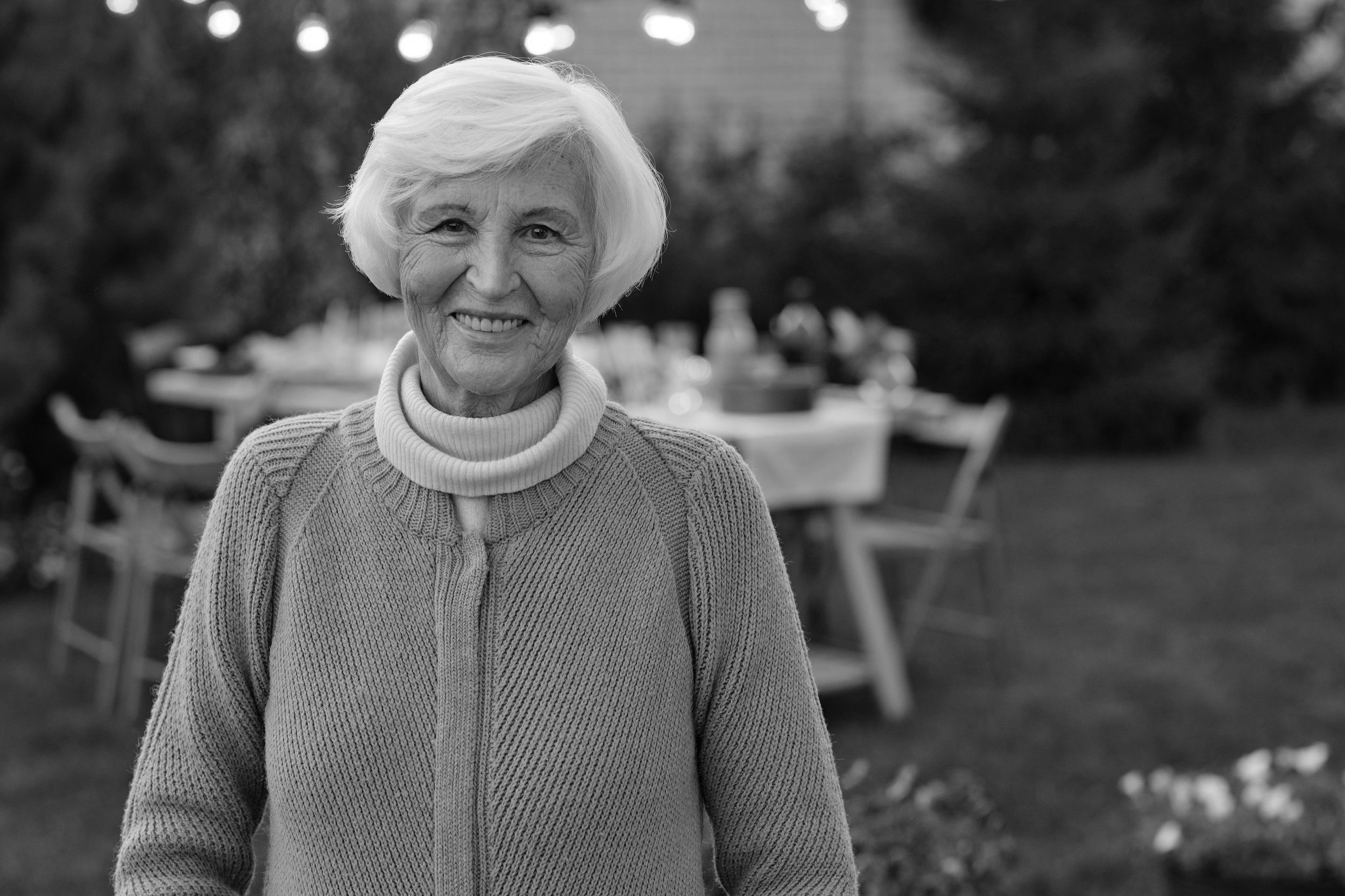 An elderly woman with short, white hair smiling outdoors, wearing a knitted sweater and a turtleneck. In the background, there is a table set for an outdoor gathering with chairs and trees.